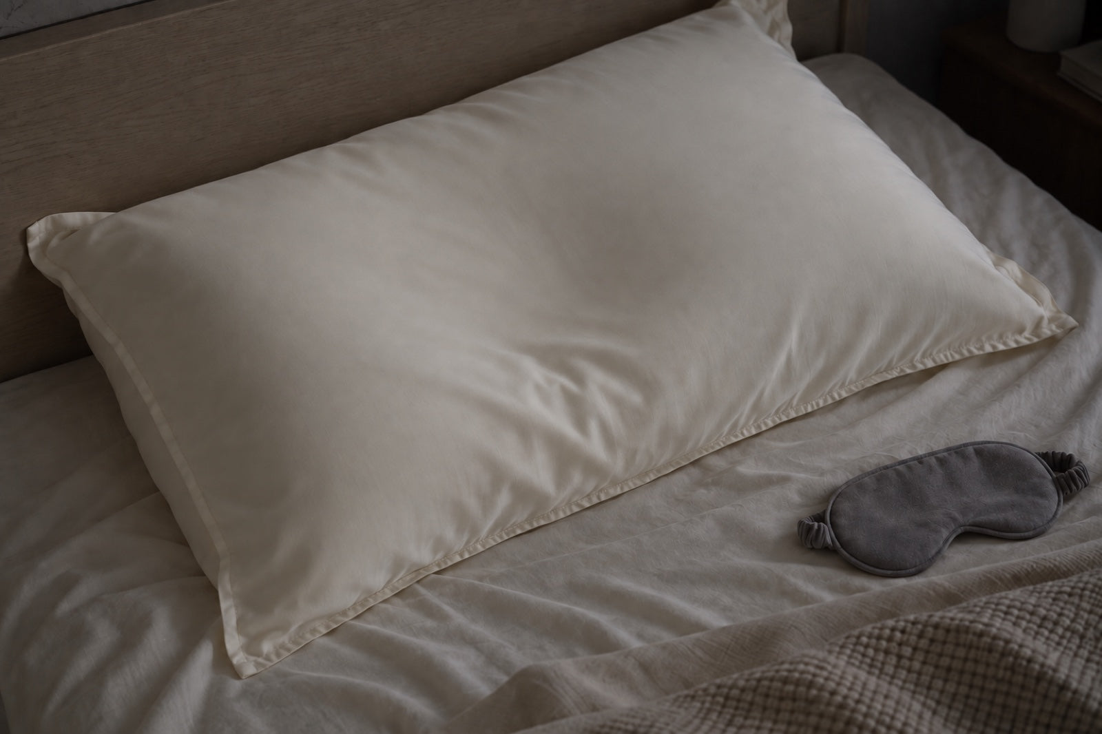 A softly settled Pearl lyocell pillow and grey eye mask in subdued nighttime light.